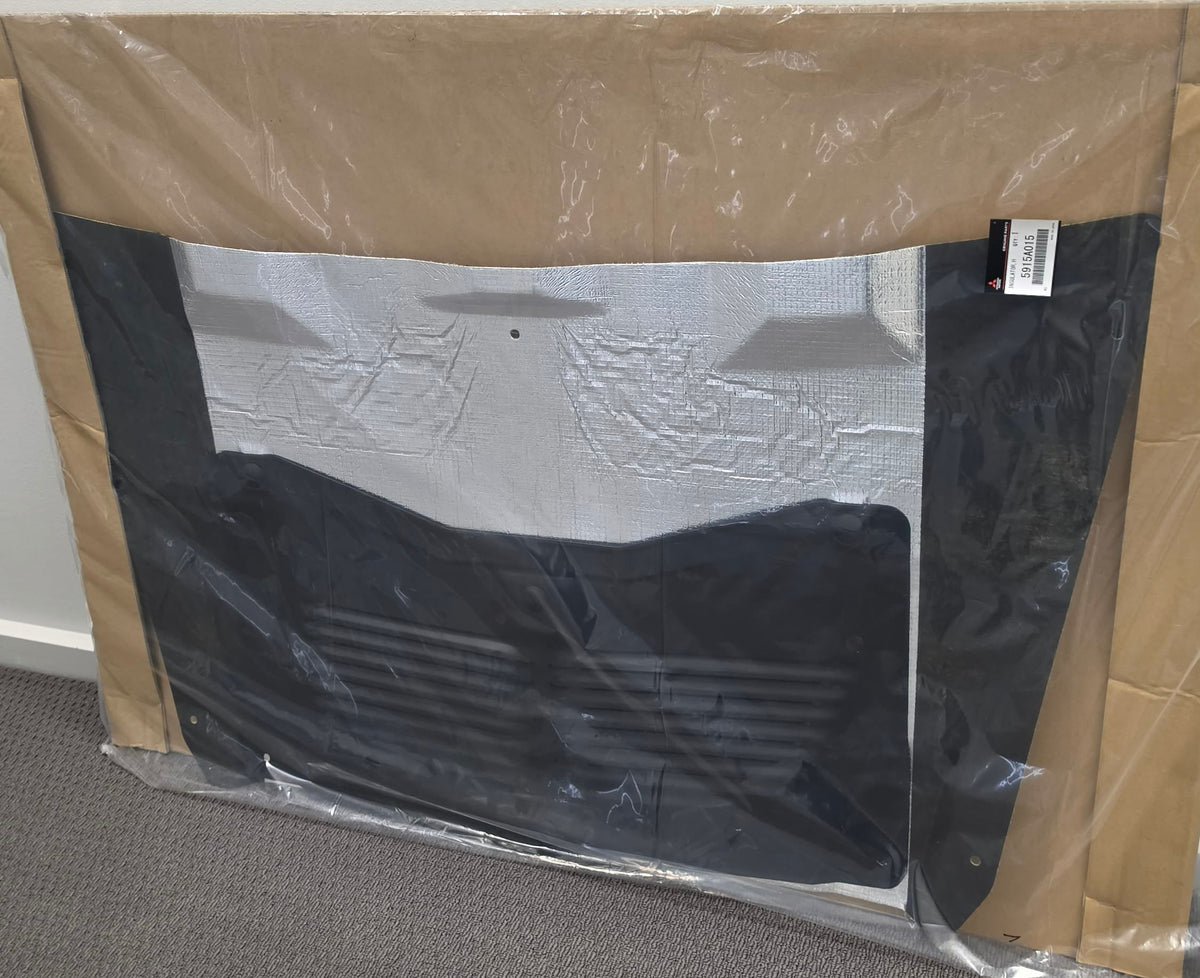 A large black car interior mat or liner wrapped in clear protective plastic, set against a brown cardboard backing on a gray carpeted floor against a white wall.