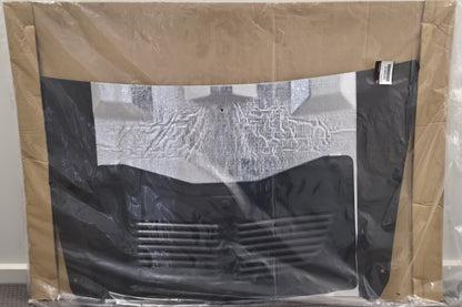 A black car grille surround with a vented lower section, wrapped in clear plastic and held in place by brown cardboard sheets, photographed against a plain indoor wall.